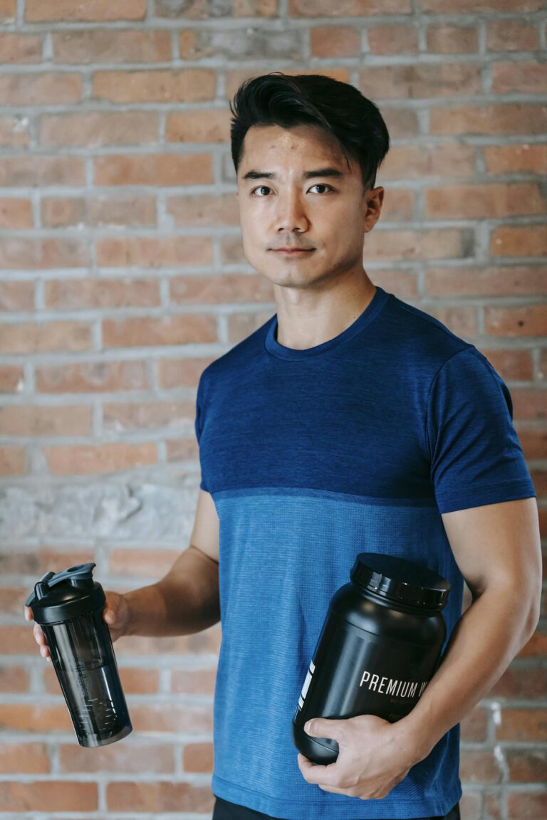 Asian man in a blue t-shirt holding two supplement bottles, standing against a brick wall.